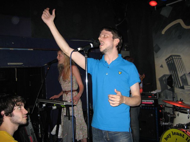 Cantante con maglietta azzurra e mano alzata vicino a ragazza che suona